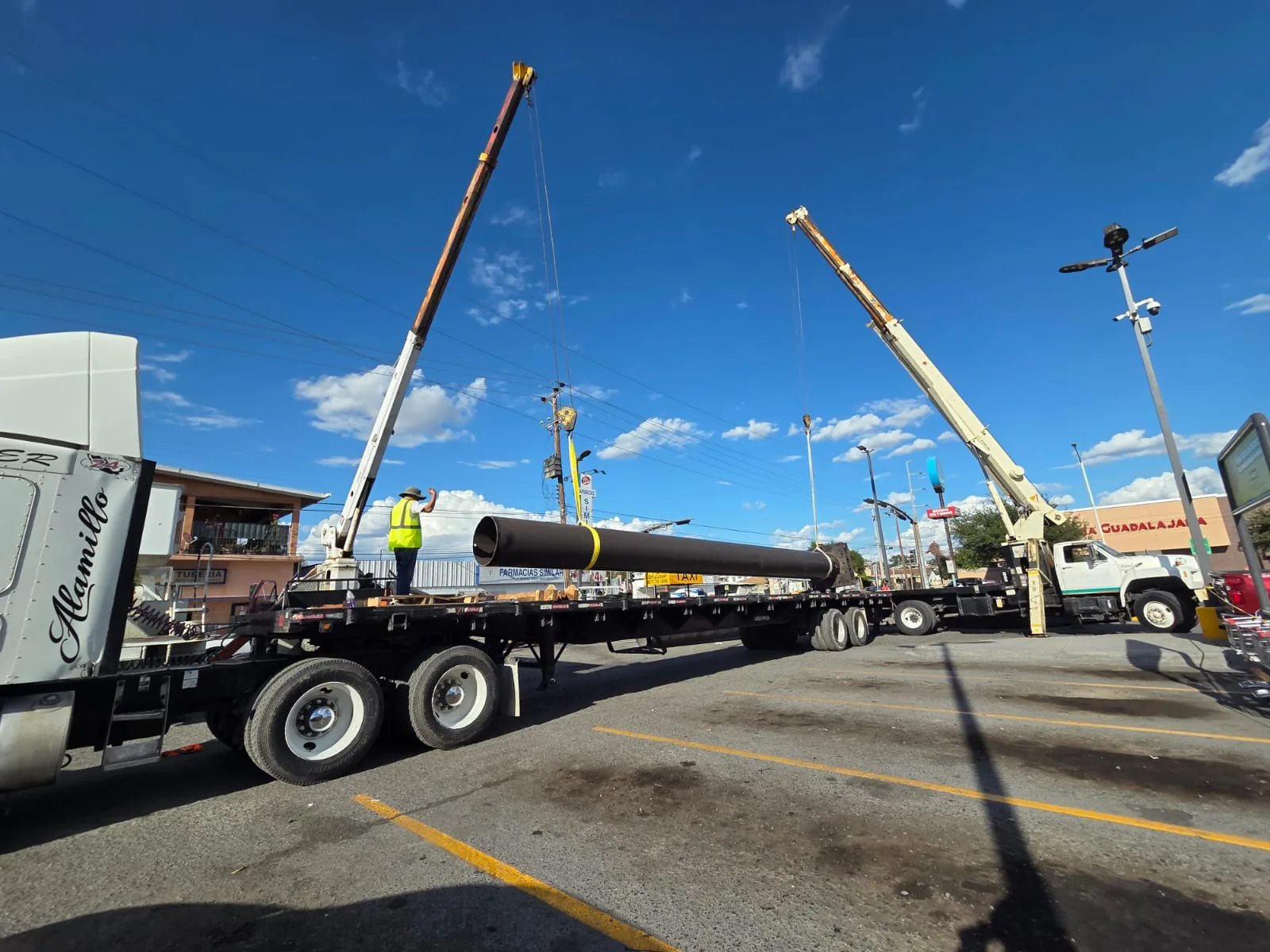 Maniobra a dos grúas para descargar un tubo de acero de una plataforma, en el estacionamiento de una plaza
